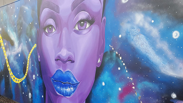 Mural of a Black woman with purple skin, blue lips against a cosmic background