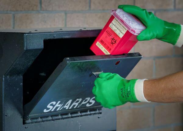 gloved hands putting a sharps container into a mailbox-sized collection bin.