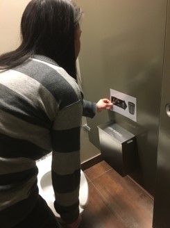 A person installs a 'Keep Trash Out of Toilets' sign in a toilet stall