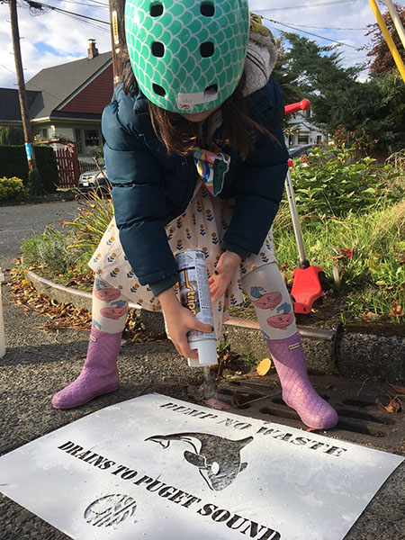 A child wearing a helmet and rubber boots stands above a storm drain and sprays white spray paint onto a stencil.