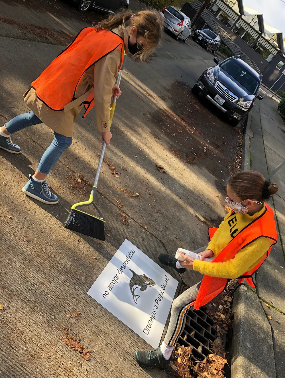 One young person wearing a safety vest sweeps the street around a storm strain while another holds a can of spray paint above a stencil reading