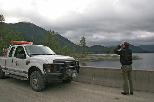 SPU employee monitoring Cedar River Watershed with binoculars, standing beside truck.