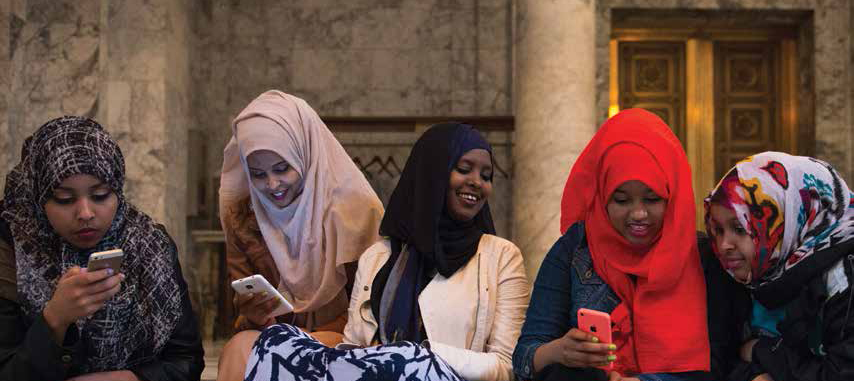 Young Somali women inside a state capitol building wearing hijabs and looking down at their phones.