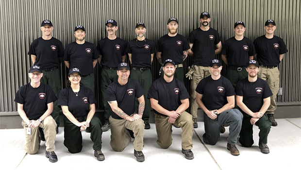 Fourteen members of the SPU Wildland Fire Crew, in matching black shirts and caps, pose for a group photo.