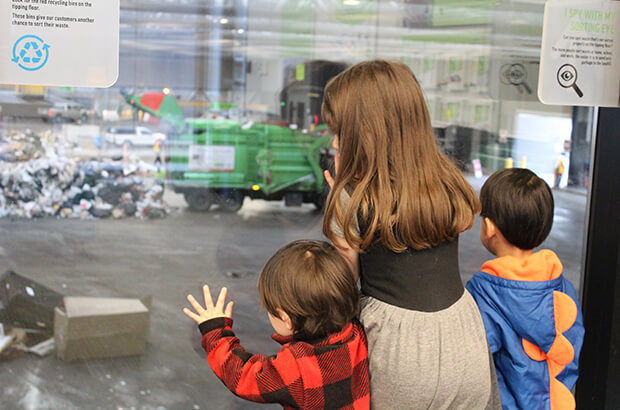 Photograph of three small children in visitors viewing room watching waste sorting operations on main floor of transfer station.