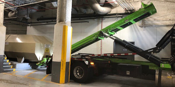 Photo shows a roll-off truck in action moving a compactor in the service area of a commercial building.