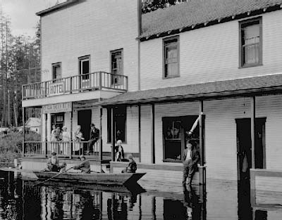 Three people in a rowboat approach the front porch of a two-story wooden hotel building in Moncton as flood waters rise to the hotel's doors.