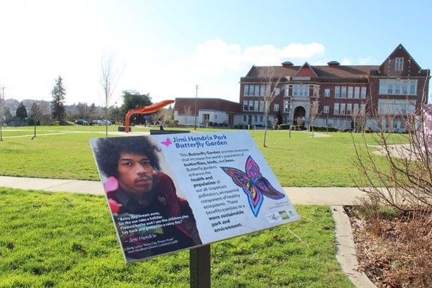 An educational sign on a post next to a garden, with a grassy field and a large building in the background