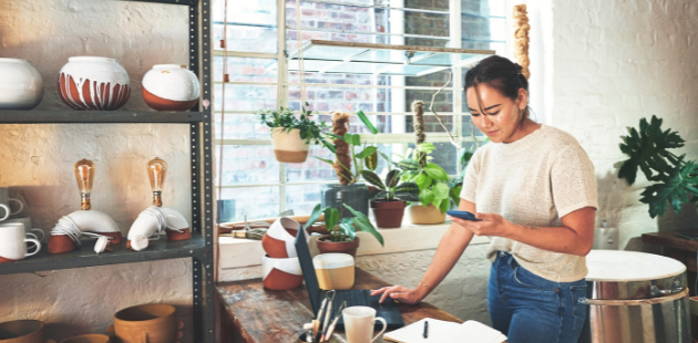 Business owner standing and using laptop and cellphone in pottery studio with ceramics and plants in background.