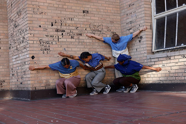 Four dancers lean against a brick wall with graffiti, their arms extended, heads down.