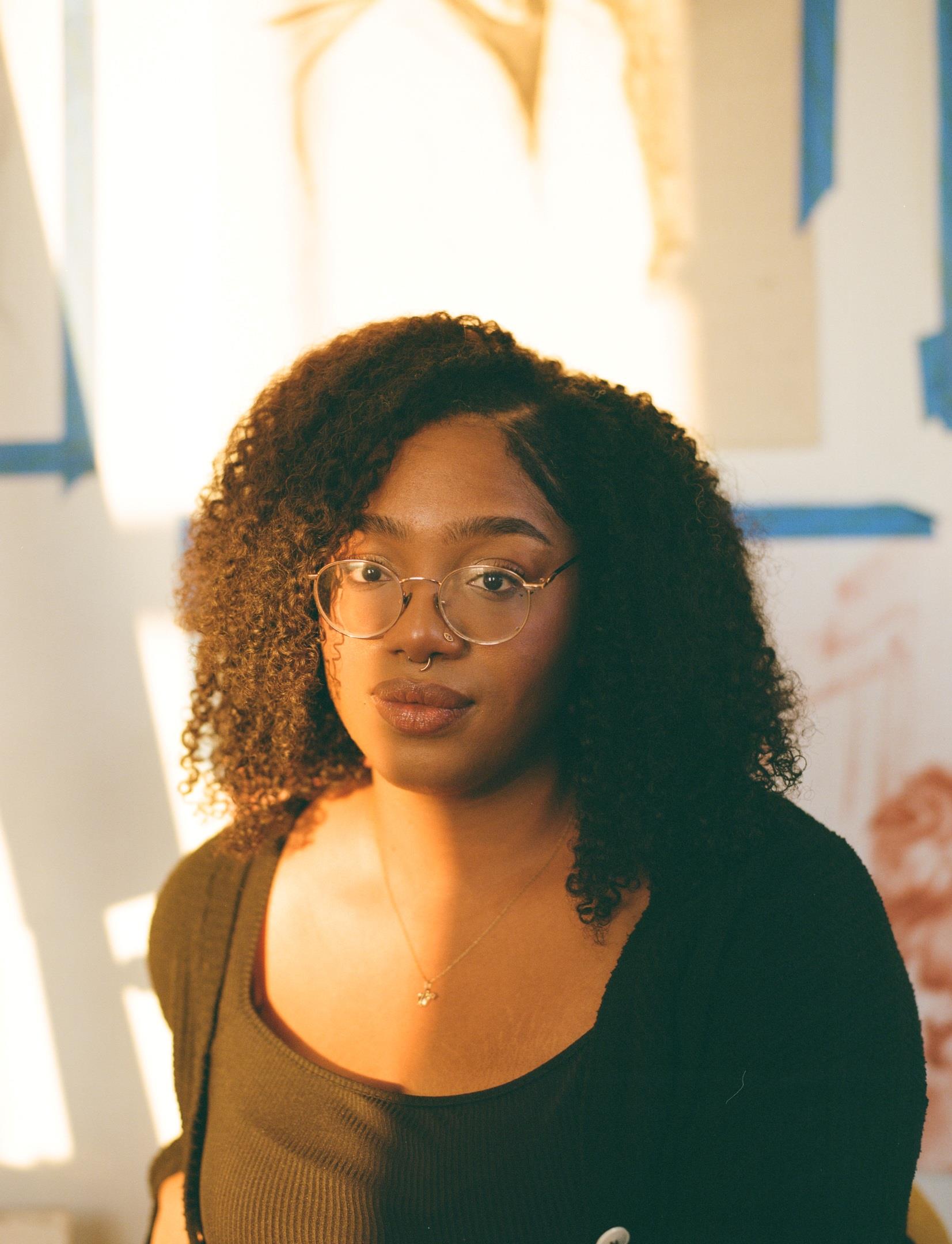 Portrait from the shoulders up of a young Black woman in a painting studio in the sunlight.