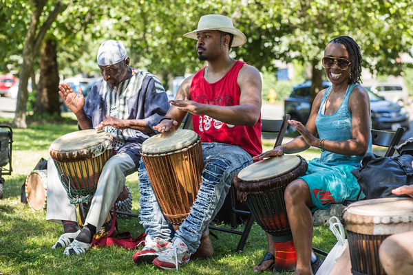 Three Black people play drums while seated in a park.