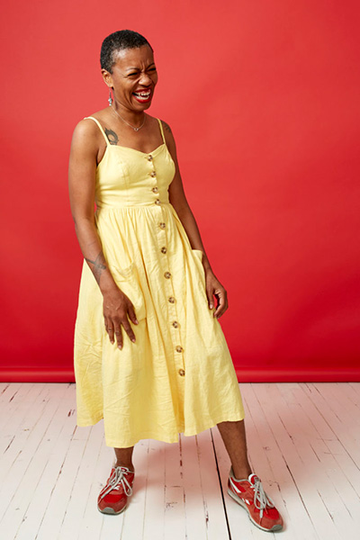 Anastacia-Renée smiling in a yellow dress and red shoes, standing in front of a red wall.