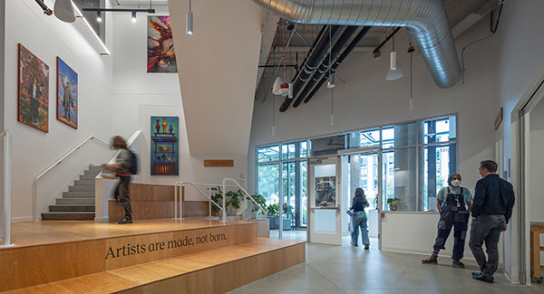 Interior art space with large windows, a wooden seating area, and a staircase.