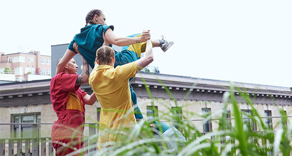 Two dancers in colorful jumpsuits hold up a third against a gray sky.