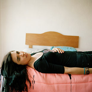 A young Latina lays across a single bed, staring straight at the viewer. Her long hair dangles off the edge of the mattress.