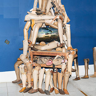 Artificial legs and suitcases arranged into a pyriamid shape against a blue wall.