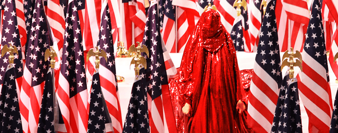 A Muslim woman in full red sparkly chador stands in front of rows of American flags.