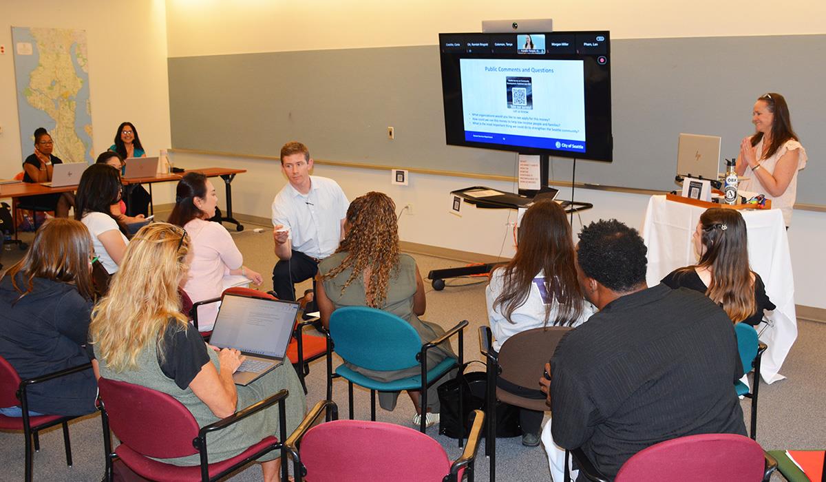 An audience member speaks into a microphone at a public hearing in front of a screen with remote participants.