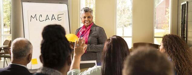 Black woman standing in front of a group of people and an easel with MCAAE written on it