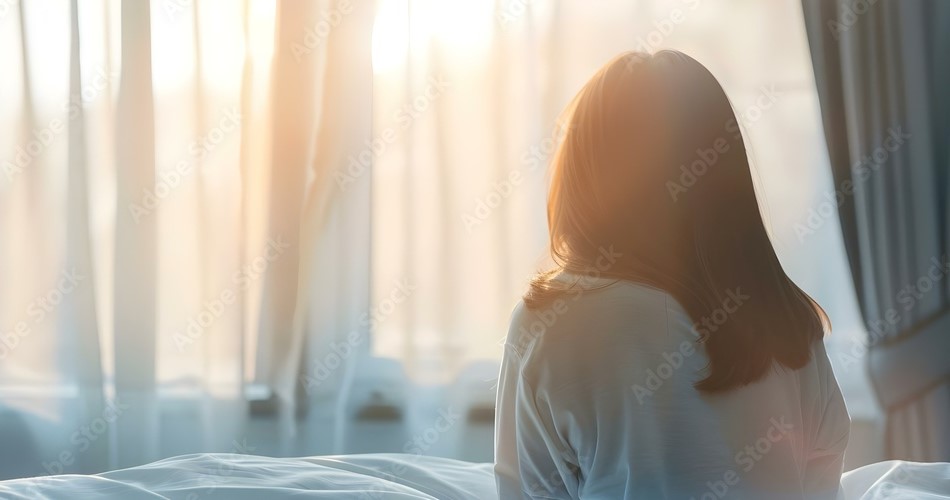 Picture of woman gazing towards a window as the sun softly shines through sheer white curtains