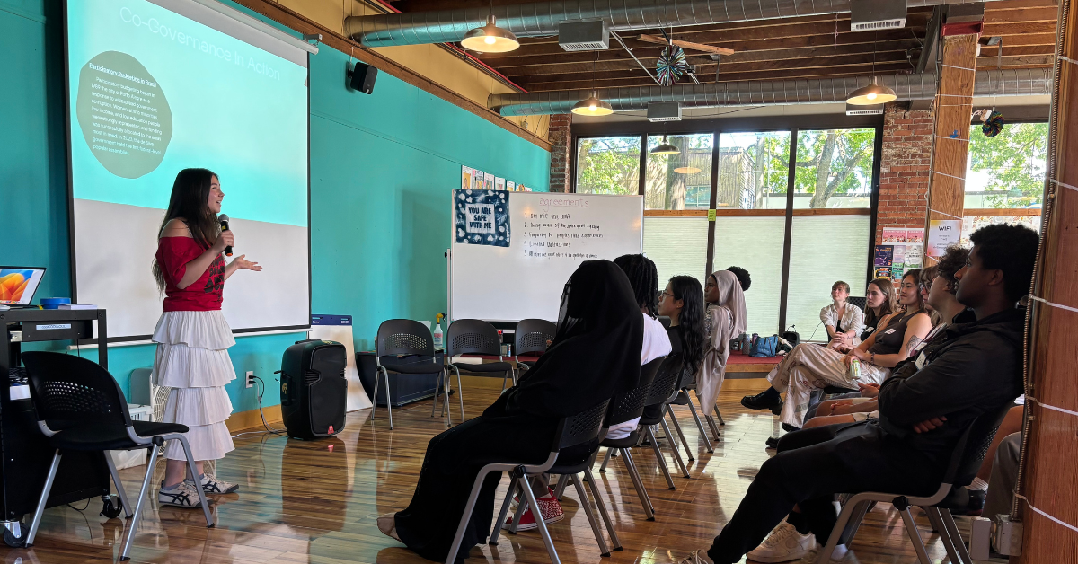  A presenter stands at the front of the room in front of two rows of participants sitting in chairs at the Youth Community Assembly