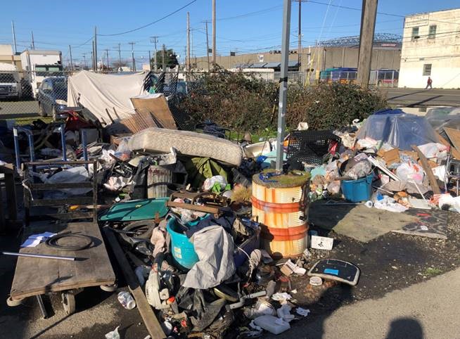 an industrial neighborhood's street corner with a 20x20 foot pile of old wet trash and plastic stacked to about 6 feet