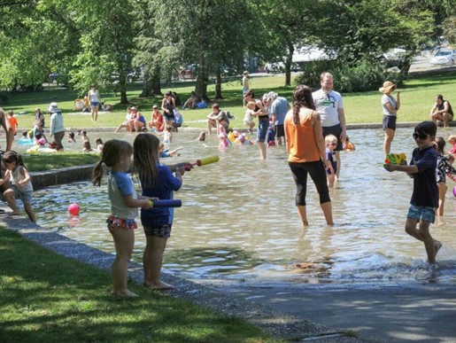 Dozens of children and their caregiver adults wade up to their ankles in a shallow pool on a sunny summer day.