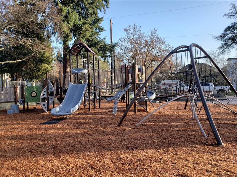 modern playground with various play structures, including multiple slides, climbing elements, and a rope dome. The ground is covered with wood chips, surrounded by trees