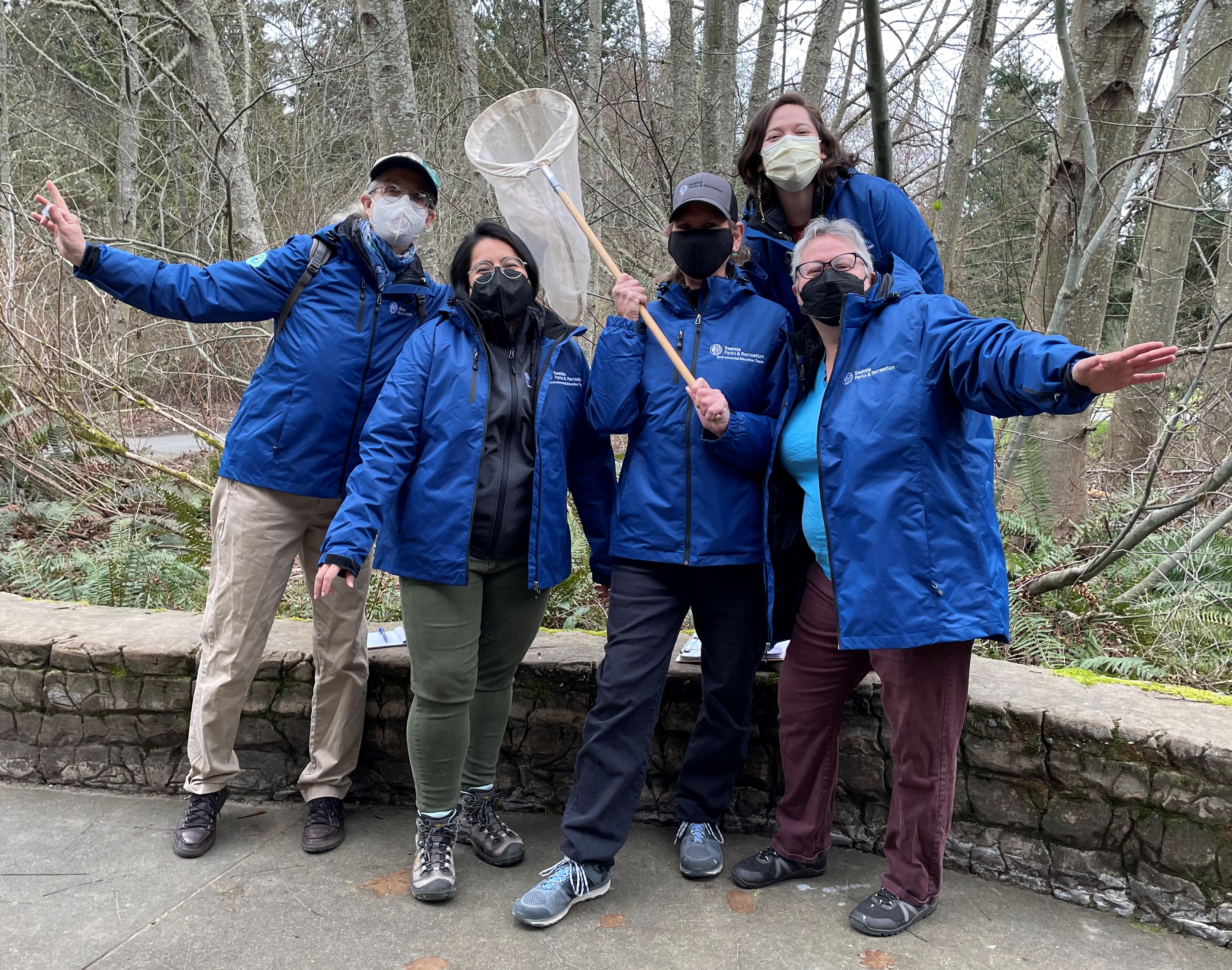 Five people in blue jackets side to side, standing outdoors in front of bare trees, smiling at camera