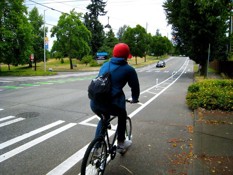 Image of a person wearing a red hat, blue sweatshirt and jeans, and a drawstring backpack biking on a protected walkway. There are paint stripes and small, white flexible delineators acting as a buffer between the bike and traffic lanes. 
