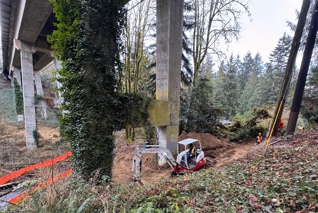 Excavator working under a bridge near a forested slope, with workers and erosion control barriers nearby.