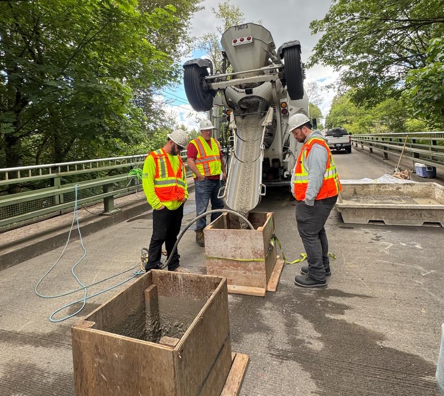 Three workers in hard hats and neon safety gear stand beside a concrete truck pouring concrete into wooden forms.