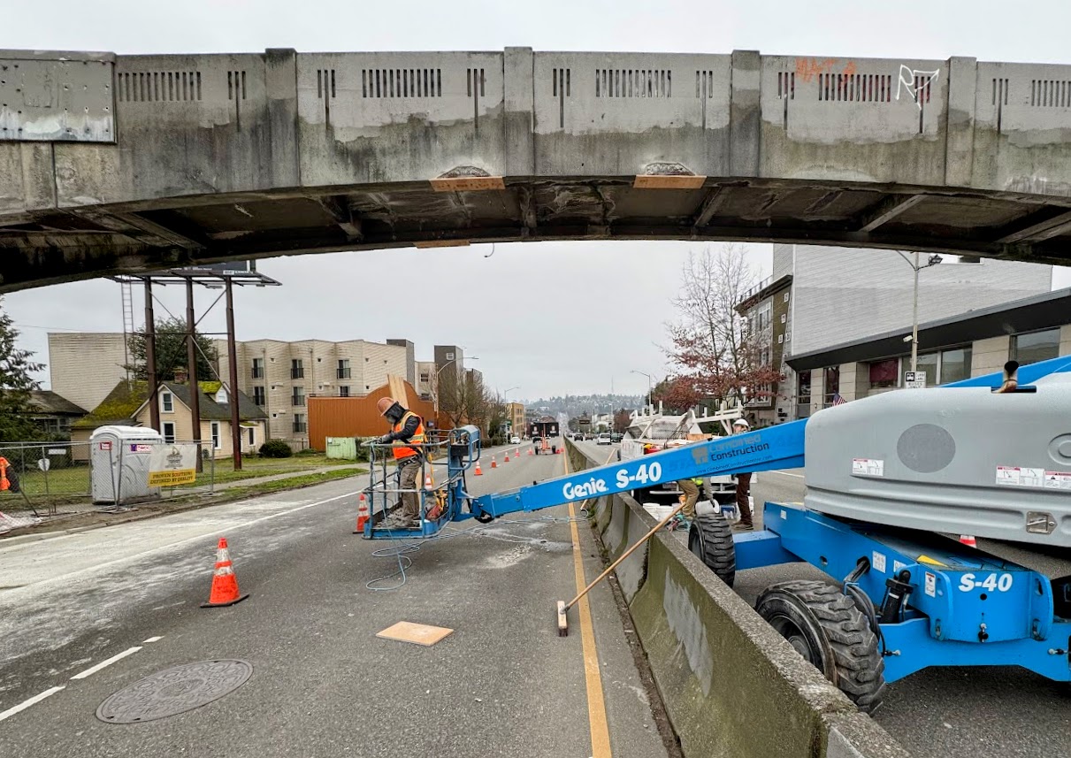 Construction workers using a lift to perform maintenance on a pedestrian bridge over a busy street. Traffic cones and barriers are in place.