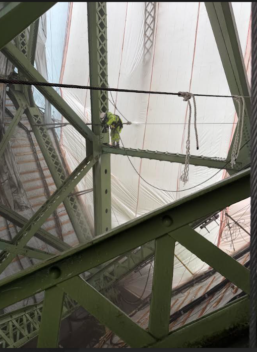 Worker in high-visibility safety gear standing on steel beams inside the structural frame of a bridge, surrounded by scaffolding and containment sheeting.