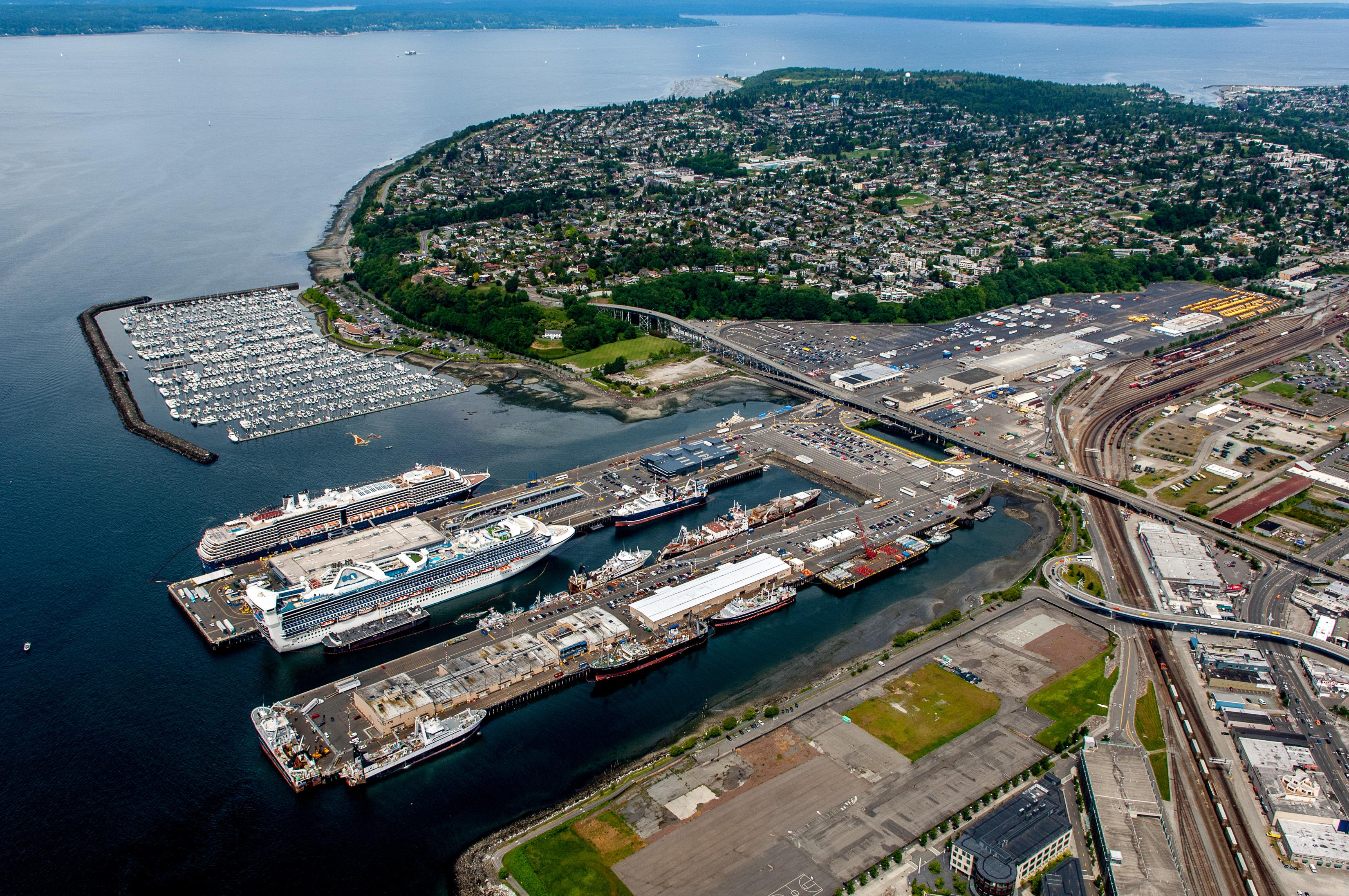 Image of the Magnolia Bridge adjacent to Terminal 91, the Smith Cove waterway, and Smith Cove marina with docked sail boats and a cruise ship. The Magnolia Bridge spans over Terminal 91 into the Magnolia neighborhood. Elliott Bay and the Olympic Peninsula are located in the background.