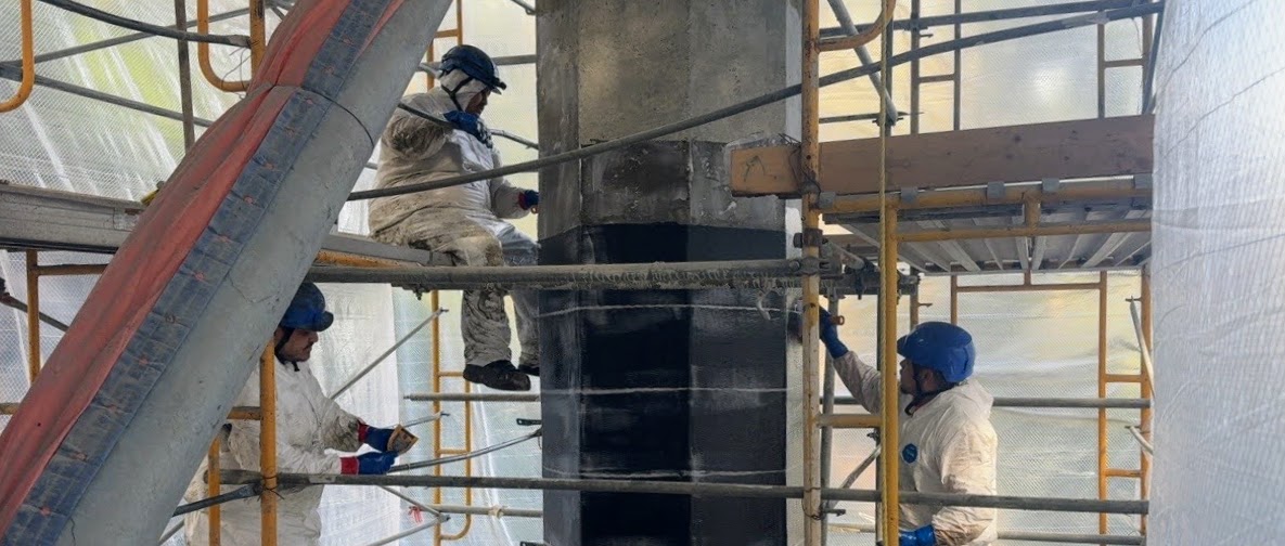 Three construction workers in safety gear apply material to a concrete bridge column surrounded by scaffolding. 