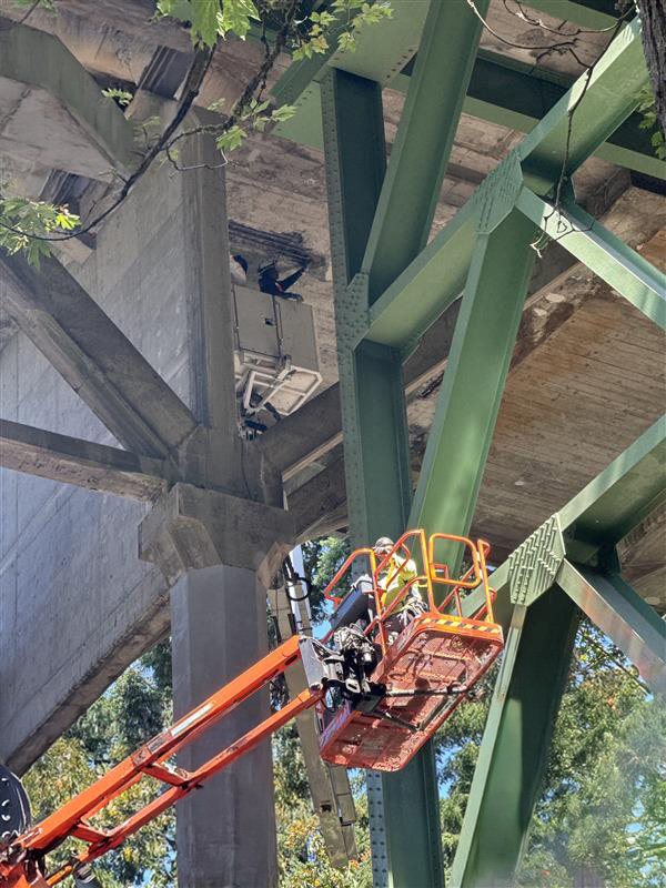 A worker in safety gear stands in an orange aerial lift beneath a bridge, repairing damaged concrete on the underside of the bridge deck.