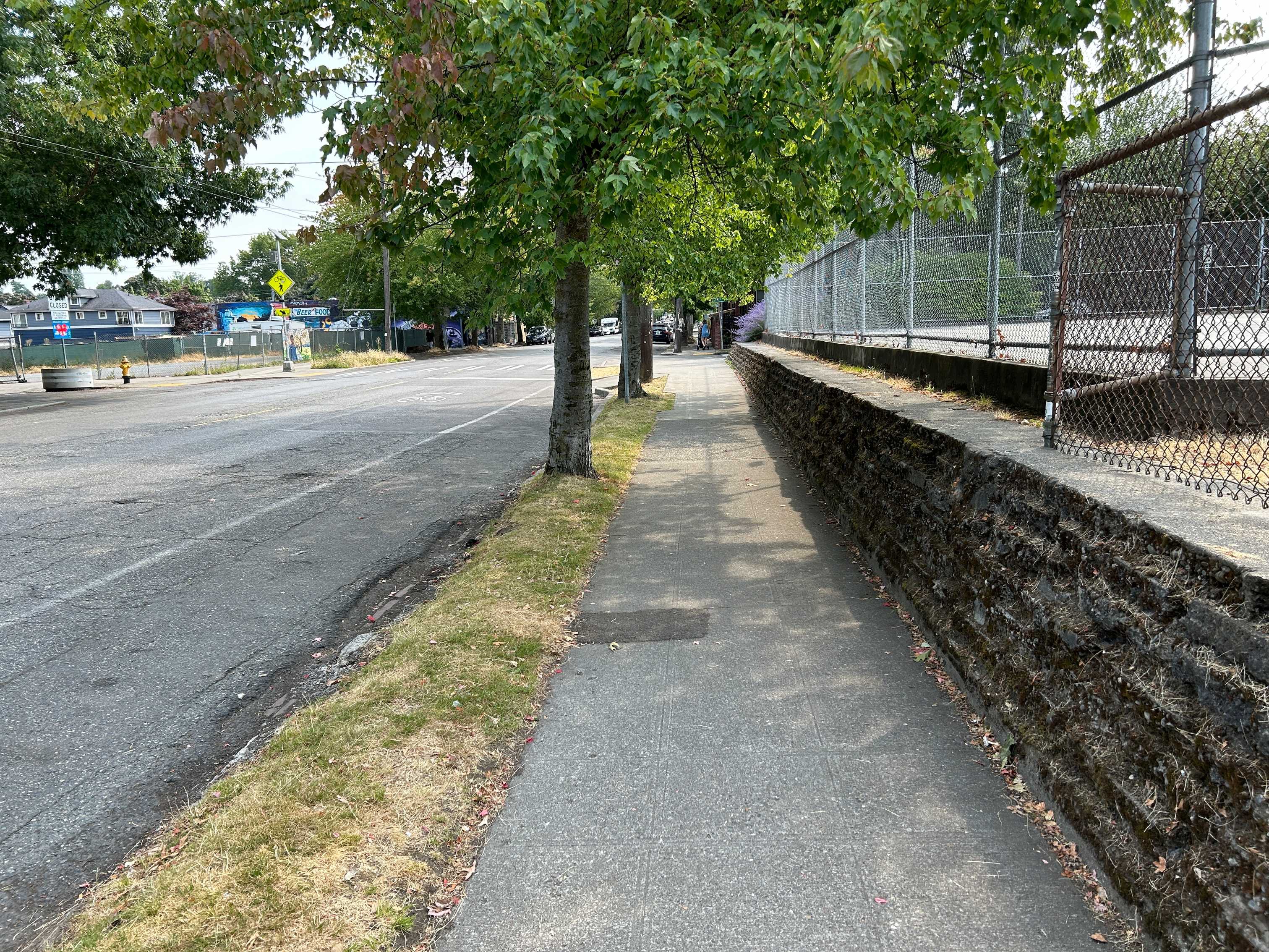 idewalk along 25th Ave with trees, a retaining wall, and a fenced sports court. The sidewalk has cracks and uneven patches. A bike lane, pedestrian crossing sign, and mural are visible in the distance.