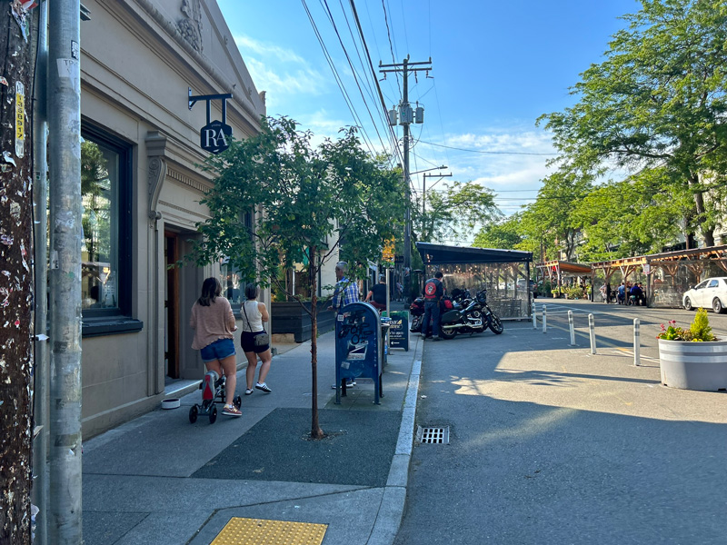 View of Ballard Ave showing cafe structures, tree pits with FPST, bollards, and planters.