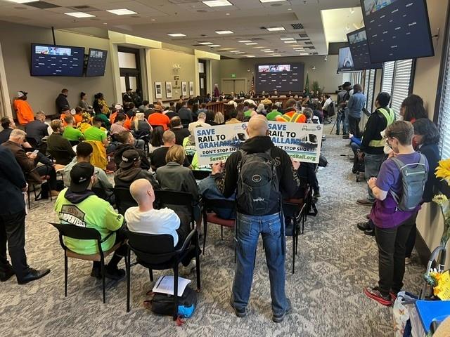 Room with about 100 people sitting and listening to people speaking at the front of the room. One person holds signs that say "Save Light Rail to Graham Street" and "Save Ballard Light Rail"