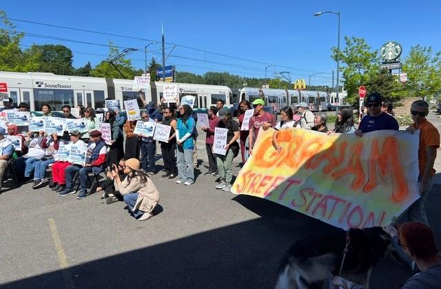A group of about 50 people stand in a semi-circle listening to a person singing and playing the guitar. Many people hold signs that say things like: "Save Graham St Station." Behind the people a light rail train passes by.