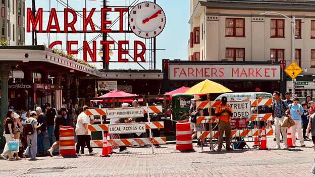 People walk under the Public Market Center sign, behind orange traffic signs that say "Street Closed, Except authorized vehicles, local access ok"