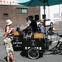 An adult and children purchasing drinks from a bicycle-drawn coffee cart with a large umbrella shading it. 