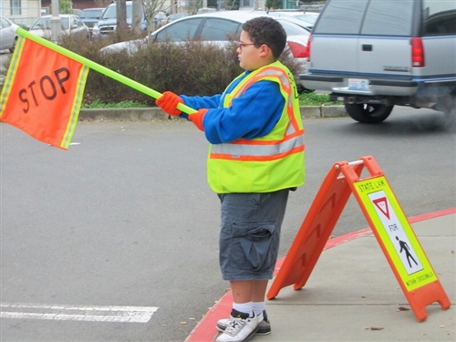 A student wearing a yellow safety vest and holding a stop flag volunteers to monitor the crosswalk at his school