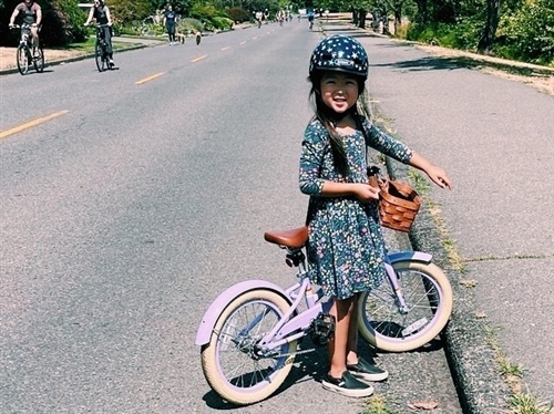 A school age child smiles with her bike on a pedestrian street.