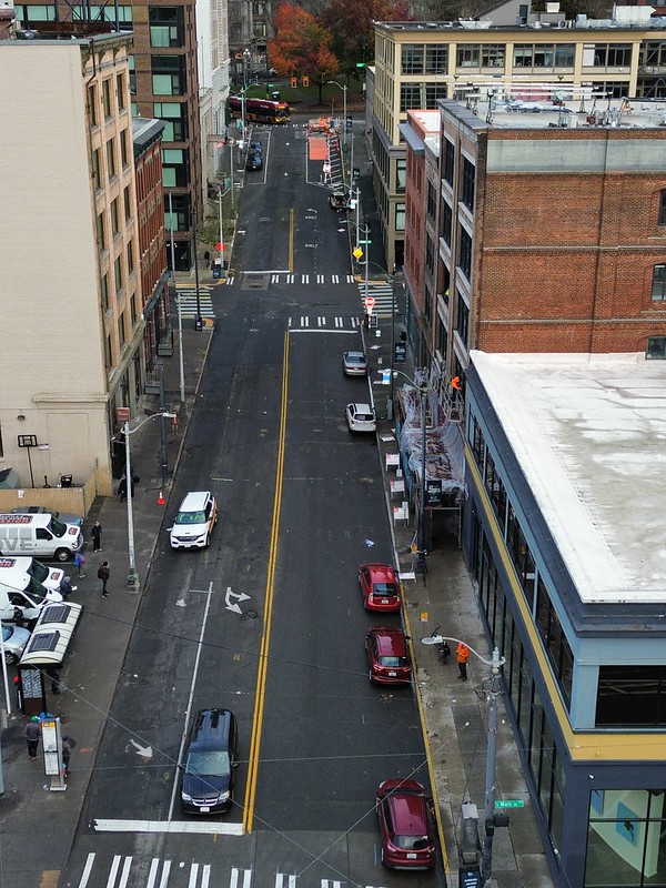 A photo taken with a drone, showing 3rd Ave facing north from S Main St to Yesler Way. Cars are parked on the east side of the street and vehicles travel southbound down the patchy, bumpy street  