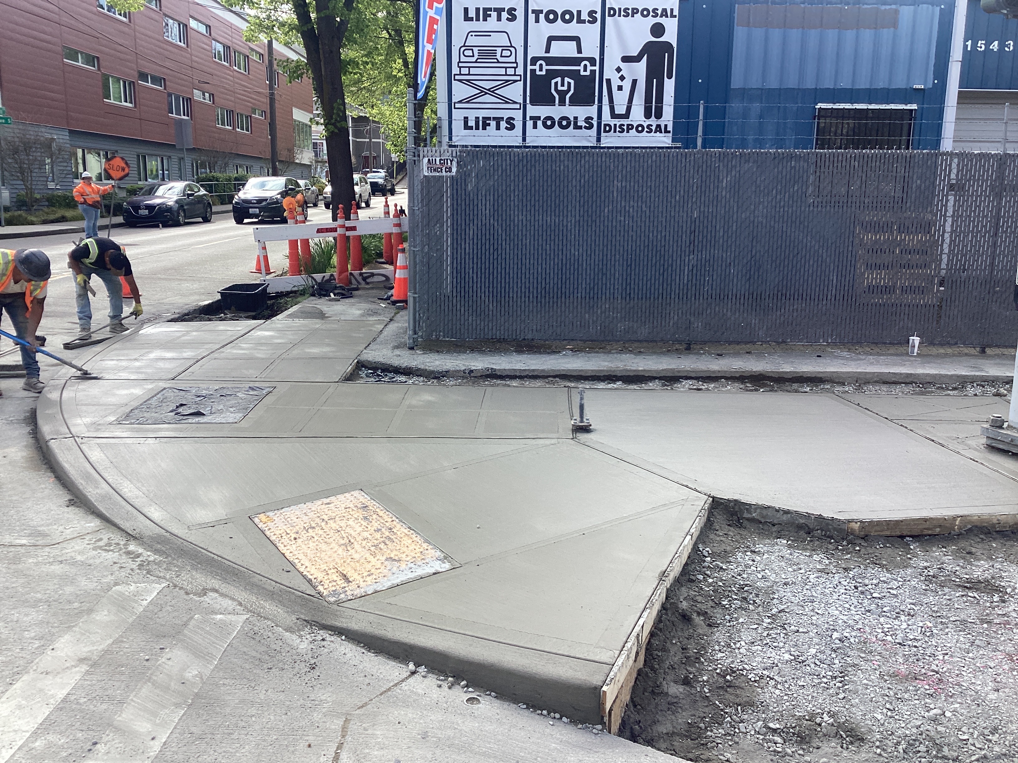 Photo showing crews finishing freshly poured concrete sidewalk and curb ramp at Rainer Ave S and S Massachusetts St.