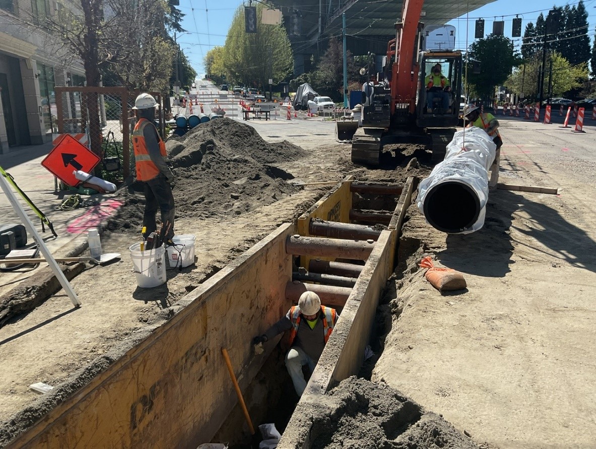 Photo of construction worker standing next to a large hole with a big pipe on the ground next to it. 