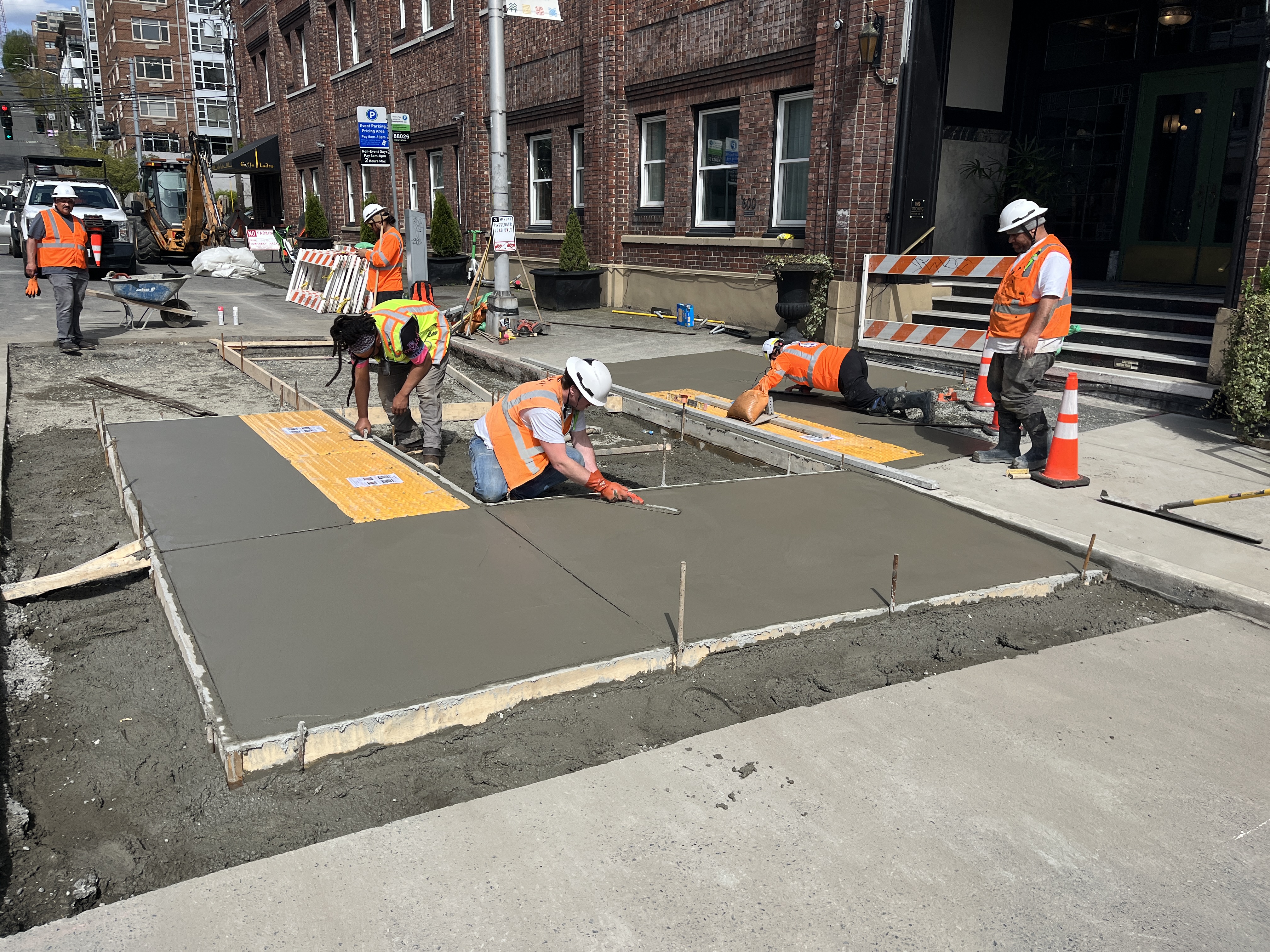 Construction workers in safety gear finish pouring and smoothing concrete panels for an ADA-compliant loading zone in front of the MarQueen Hotel on Queen Anne Ave N.